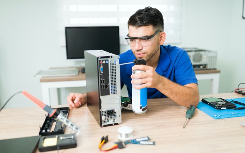 Dirty computer. Attractive young man wearing protection glasses while cleaning inside with compressed air the hardware of a computer