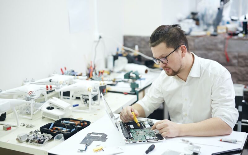 Portrait of modern man wearing glasses busy working with electronics in workshop: disassembling laptop and drones with different tools and electronic devices on table
Keywords: